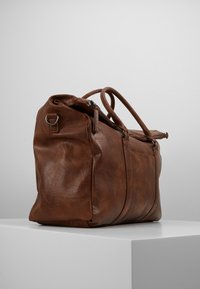 Braune Lederreisetasche mit zwei oberen Griffen, strukturiertem Oberflächen, Reißverschlussverschluss, seitlicher Tasche und Metallakzenten.