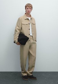 Giacca beige con due tasche a pattina, abbinata a pantaloni coordinati. Una borsa nera è tenuta in una mano; le scarpe scure con lacci completano l'outfit.