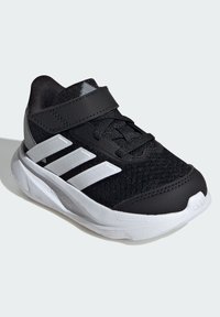 Scarpa da atletica nera con tomaia in mesh, cinturino regolabile e design a tre strisce bianche. Presenta una suola esterna bianca testurizzata e un colletto imbottito.