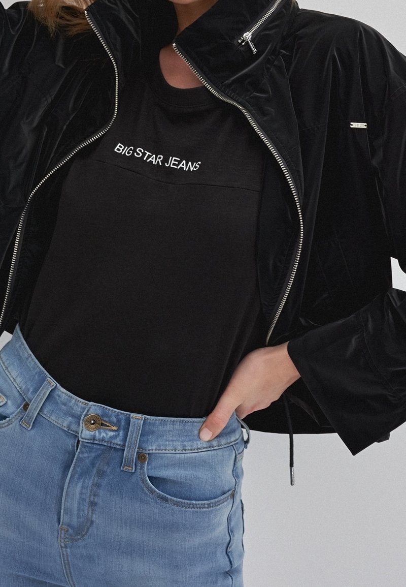 Person som bär en svart t-shirt med texten "BIG STAR JEANS", en svart jacka med dragkedja och ljusblå jeans med en hand i fickan.