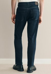 Tumed sinised denim teksad otse lõikega, kahe tagatasku ja peene tekstuuriga. Kanda mustade kingadega, neutraalse tausta taustal.