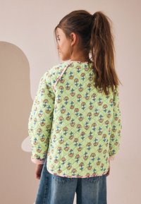 Veste matelassée verte avec un motif floral jaune et bleu, bordée d'un galon rose le long de l'encolure et des poignets. Vue arrière présentée.