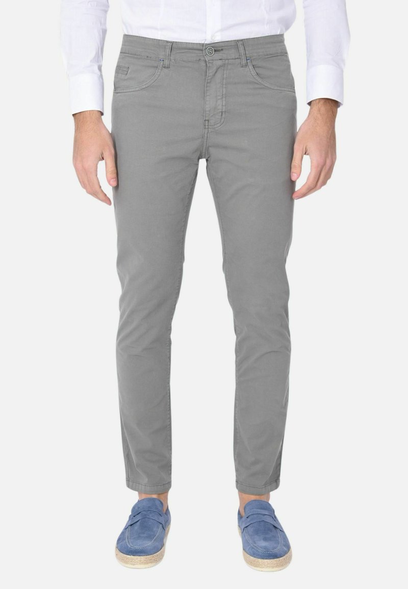 Chinos grigio chiaro, slim-fit, con cinque tasche, caratterizzati da una texture liscia e dettagli di cucitura sottili. Indossati con scarpe blu slip-on.