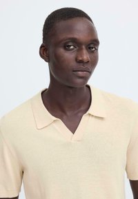 Polo en maille beige avec col en V, manches courtes et texture lisse. Présente un col et des poignets côtelés avec un design minimaliste.