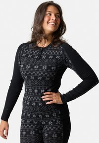 Ung kvinde iført sort langærmet top og leggings med grå snefnug- og diamantmønstre, smilende og kigger til siden.