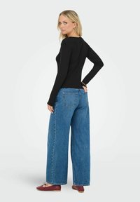Czarna bluzka z długim rękawem, w prążki, w połączeniu z niebieskimi, szerokimi dżinsami, z falbaniastym dołem. Modelka ma na sobie bordowe baleriny.