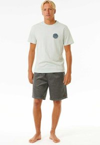 Lichtblauw t-shirt met korte mouwen en een ronde graphic op de borst, gecombineerd met donkergrijze knie-lange shorts, model staat blootsvoets.