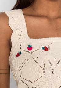 Krémový pletený top bez rukávů s volánovými ramínky a malou vyšitou červenou jahodou na oblasti hrudníku.