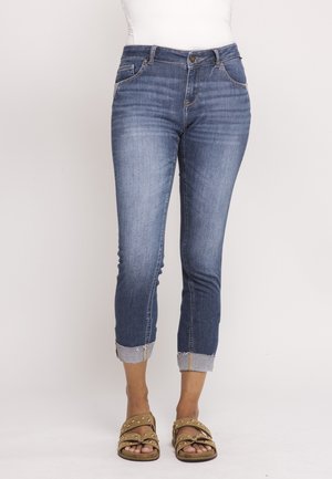 Woman wearing blue rolled-up skinny jeans and beige studded sandals, standing against a plain light background.