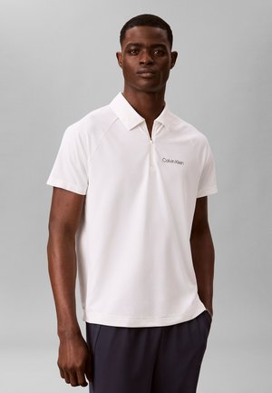 Uomo con pelle scura che indossa una polo bianca Calvin Klein a maniche corte e pantaloni scuri, in piedi davanti a uno sfondo grigio uniforme.