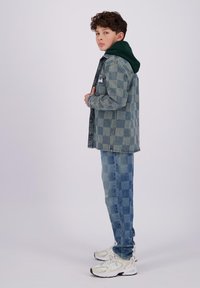 Giacca di jeans con un motivo a scacchi, abbinata a un felpa verde e jeans a scacchi blu. Sneakers bianche con dettagli.