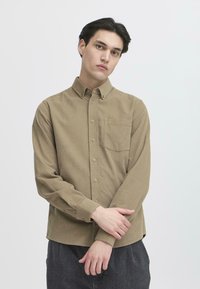 Giovane uomo con capelli scuri indossa una camicia button-up beige chiaro, in piedi con le braccia incrociate su uno sfondo bianco semplice.