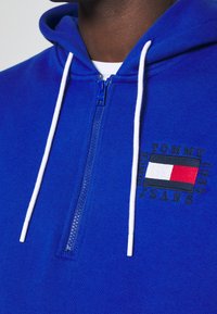 Felpa con zip blu realizzata in tessuto morbido, con stringhe bianche e un logo ricamato in rosso, bianco e blu sul petto.