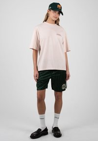 Femme portant un t-shirt beige oversize, un short rayé vert foncé, des mocassins noirs, des chaussettes blanches et une casquette de baseball noire avec un logo NY orange.