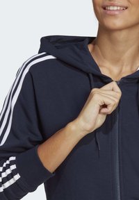 Person som ler medan de delvis drar upp en marinblå hoodie med vita ränder på ärmarna.