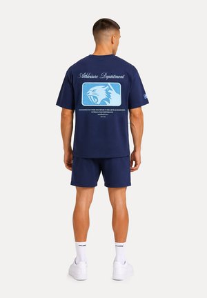 Mann steht mit dem Rücken zur Kamera, trägt dunkelblaue Sportshorts und ein T-Shirt mit einem blauen und weißen Wildcat-Logo und Text auf dem Rücken.