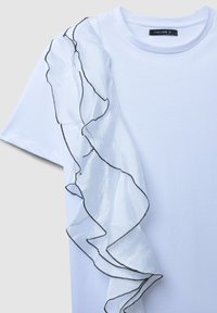 Camicetta bianca a maniche corte con tessuto trasparente e arricciato che si sviluppa diagonalmente sulla parte anteriore, rifinita con cuciture nere, scollatura rotonda.