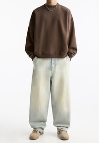 Felpa marrone con una vestibilità rilassata, abbinata a jeans a gamba larga di un blu chiaro. Le sneakers sono beige con dettagli marroni. Design semplice e coerente.