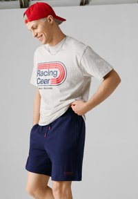 Νέος άνδρας που φοράει κόκκινο καπέλο, ανοιχτό γκρι μπλουζάκι με την επιγραφή "Racing Gear" και σκούρο μπλε σορτς, στέκεται χαλαρός μπροστά σε απλό φόντο.