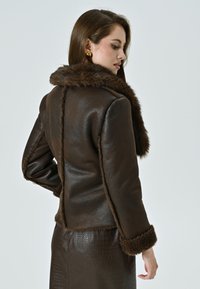 Giacca in finta pelle marrone con un morbido colletto e polsini in pelliccia, caratterizzata da una silhoutte aderente e cuciture visibili lungo il retro.
