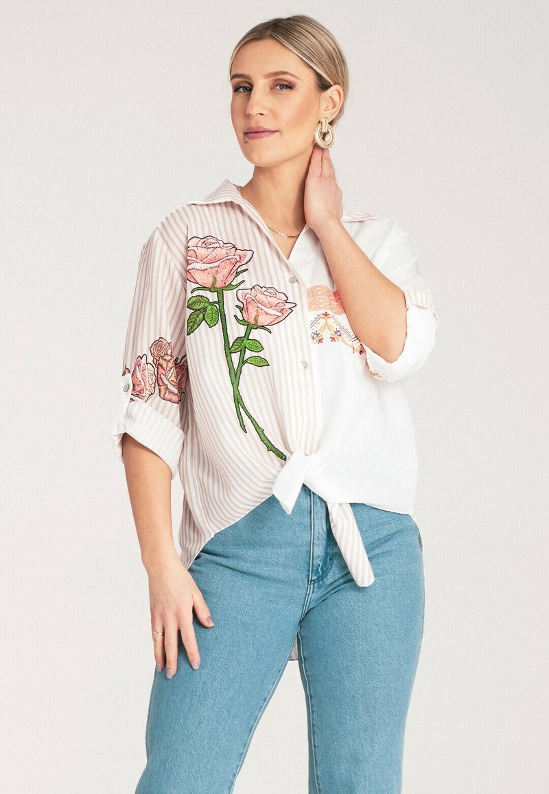 Biała i różowa koszula w paski z kwiecistymi haftami, krótkie rękawy i zawiązany przód; zestawiona z niebieskimi dżinsami o wysokim stanie.