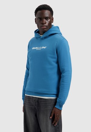 Man in een blauwe hoodie met de tekst "BALLIN'", staand met een serieuze uitdrukking tegen een effen lichtgrijze achtergrond.