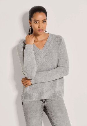 Woman with dark hair pulled back wearing silver drop earrings, light grey V-neck knit sweater, and light grey jeans, standing with one hand near face.