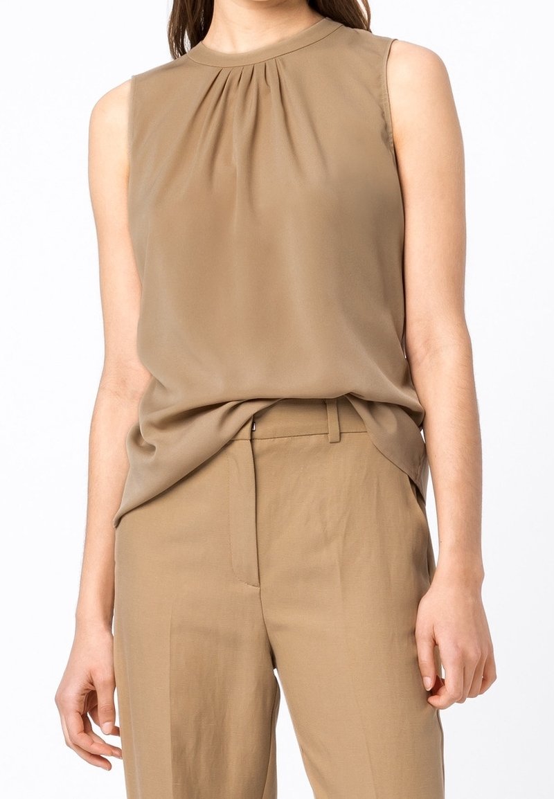 Femme portant un chemisier sans manches marron clair rentré dans un pantalon taille haute assorti sur fond blanc.