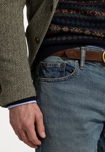 Herringbone patterned blazer in beige and black, layered over a multicolored knitted sweater. Light blue shirt cuff visible. Blue jeans with brown belt.