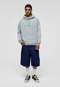 Grijze sweatshirt met spetterpatronen, kangoeroezak, gecombineerd met losse blauwe denim shorts. Zwarte sneakers, witte sokken, zwarte pet.