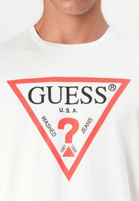 Balts kokvilnas T-krekls ar lielu sarkanu trīsstūri iezīmētu logo, kurā ir teksts "GUESS," "U.S.A.," un "WASHED JEANS", kā arī centrālais jautājuma zīmes dizains.