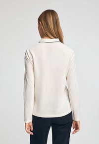Camicia bianca a maniche lunghe con colletto e dettagli in blu navy, realizzata in un materiale morbido, con un taglio dritto e un design minimalista.