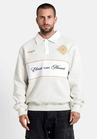 Witte sweatshirt met een polokraag, voorzien van gouden borduring, een horizontale tekstband met de tekst "Elevate Every Moment" en geribde manchetten.