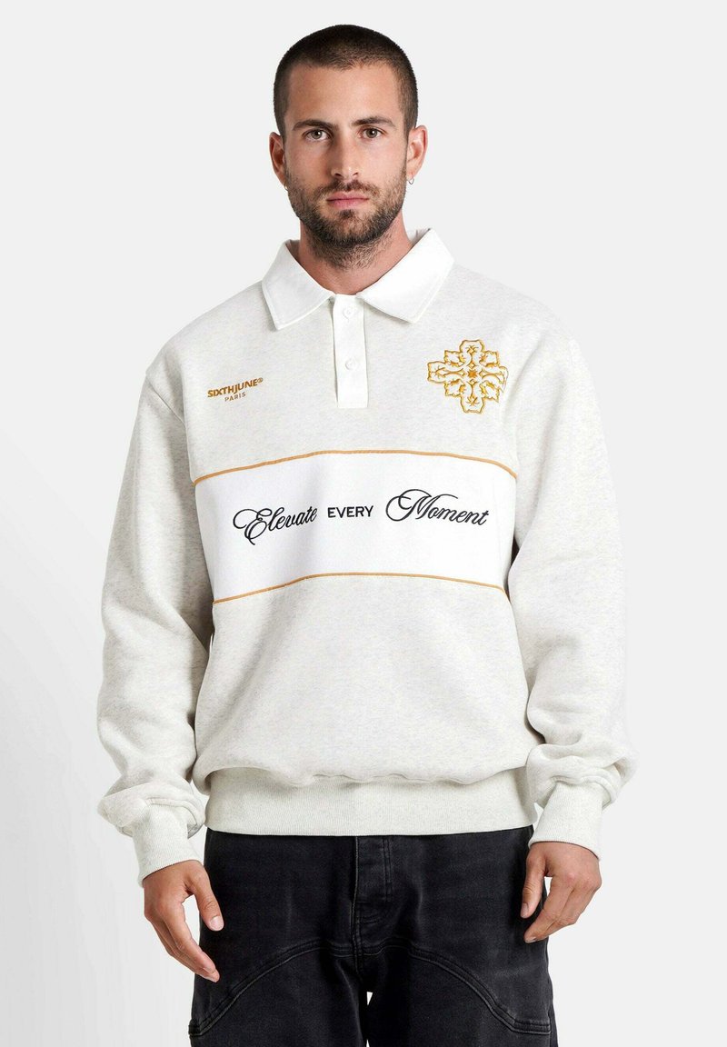 Witte sweatshirt met een polokraag, voorzien van gouden borduring, een horizontale tekstband met de tekst "Elevate Every Moment" en geribde manchetten.