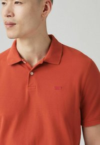 Orange bomullspolo med krage, två knappar och en liten röd logotyp på bröstet. Har en mjuk yta och korta ärmar.