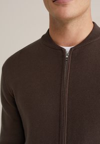 Férfi, barna kötött cipzáras pulóvert visel fehér ing fölött, amelyen a mellkas és a nyak területe látható, egyszínű háttér előtt.