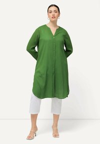 Green button-up tunic with a V-neck, long sleeves, and a relaxed fit. Paired with white cropped pants and strappy heeled sandals.