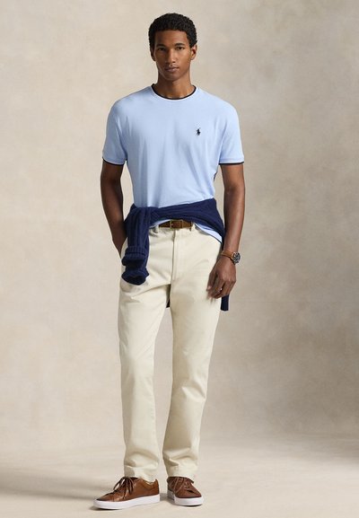 Ανοιχτό μπλε κοντομάνικο πουκάμισο με σκούρο μπλε γιακά, σε συνδυασμό με κρεμ παντελόνια και καφέ παπούτσια. Σκούρο μπλε πουλόβερ δεμένο γύρω από τη μέση. Ορατό καφέ ρολόι.