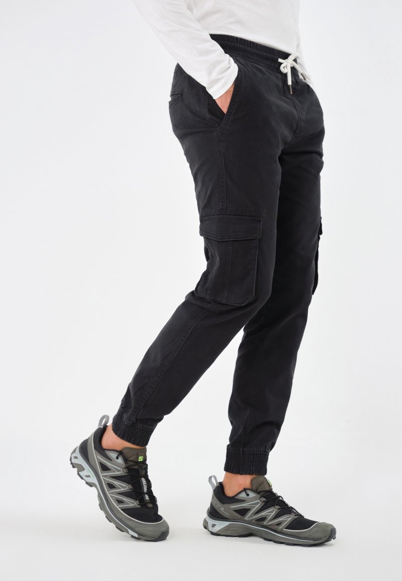 Pantalon cargo noir avec poignets élastiques et poches, associé à des chaussures de sport grises et noires avec une semelle texturée et un système de laçage.