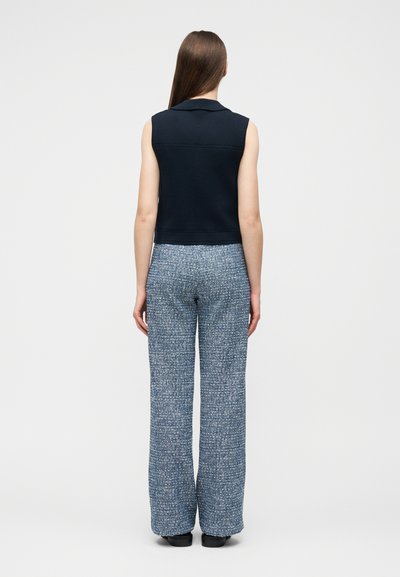 Γυναίκα που στέκεται πλάτη, ντυμένη με μαύρο αμάνικο τοπ και μπλε υφασμάτινη παντελόνα με φαρδιά πόδια, μπροστά σε απλό φωτεινό φόντο.