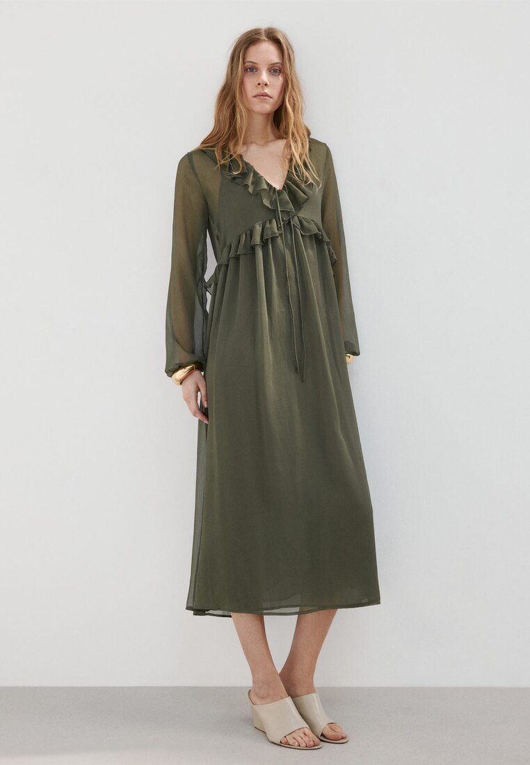 Femme portant une robe midi olive à manches longues avec volants et des sandales compensées beiges à bout ouvert, debout devant un fond uni.