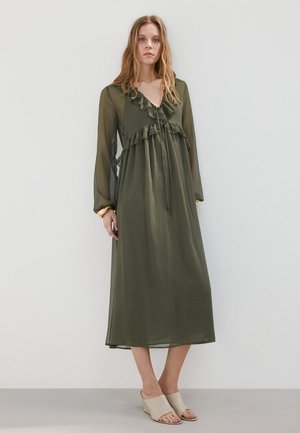 Donna che indossa un abito midi verde oliva a maniche lunghe con volant e sandali beige con zeppa e punta aperta, in piedi davanti a uno sfondo semplice.