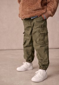 Olivgrön cargobyxor i bomull med elastiska mudd och sidofickor, kombinerade med vita sneakers. En del av en texturerad brun fleece-tröja är synlig.