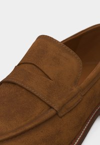 Καφετί μοκασίνι από σουέντ με διακοσμητικό λουράκι μπροστά και λεπτομερείς ραφές κατά μήκος των άκρων.