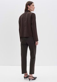 Donna in piedi di lato che indossa un top a maniche lunghe marrone scuro, pantaloni coordinati scuri e scarpe con tacco basso slingback nere su uno sfondo bianco.