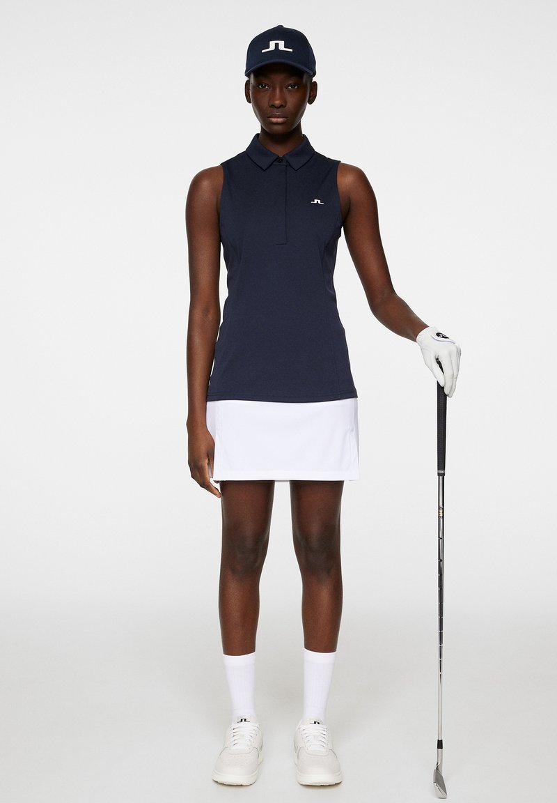 Polo senza maniche blu navy con colletto, gonna corta bianca, calze bianche fino al ginocchio e sneakers bianche. Tiene in mano una mazza da golf e indossa un cappellino blu navy.