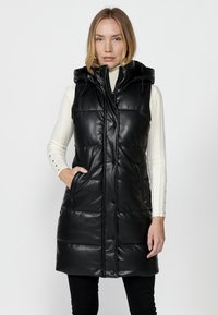 Čierny puff vest s kapucňou, vyrobený z lesklého syntetického koženého materiálu, s predným zipsom a cvokmi, spolu so bočnými vreckami.