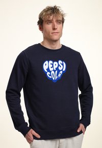 Námořnická modrá mikina s kruhovým výstřihem vyrobená z měkkého materiálu, na přední straně s velkým bílým a modrým logem "PEPSI COLA". Klasický střih.