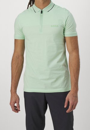 Mann trägt ein hellgrünes, kurzärmeliges Poloshirt mit Reissverschlusskragen und dunkle Hosen und steht vor einem schlichten Hintergrund.