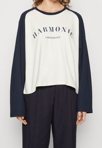 Langärmliges Raglan-T-Shirt in Creme und Marineblau. Mit "HARMONIC" und "COEXISTENCE" in Dunkelblau auf der Brust bedruckt. Aus weichem Baumwollmix.